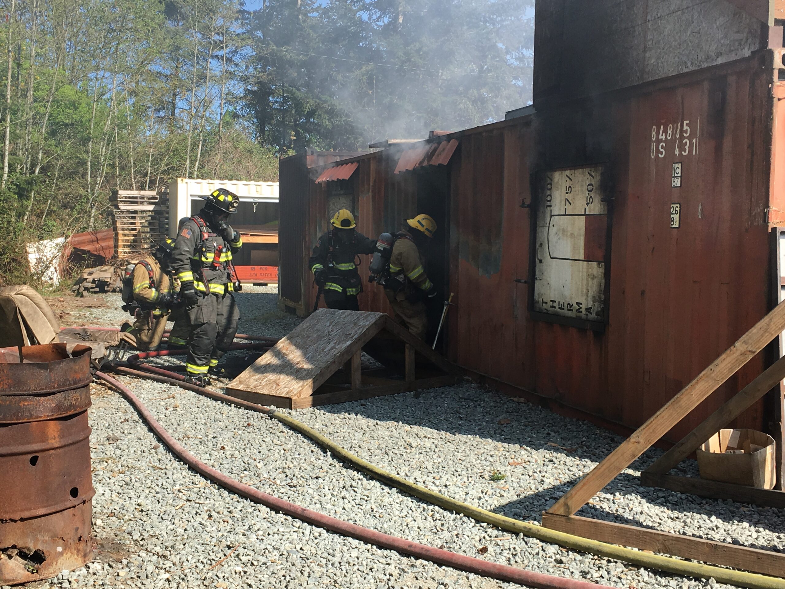 Firefighters in bunker gear and yellow helmets enter a rust red shipping container. Smoke billows from the rear of the container. It is a training prop.