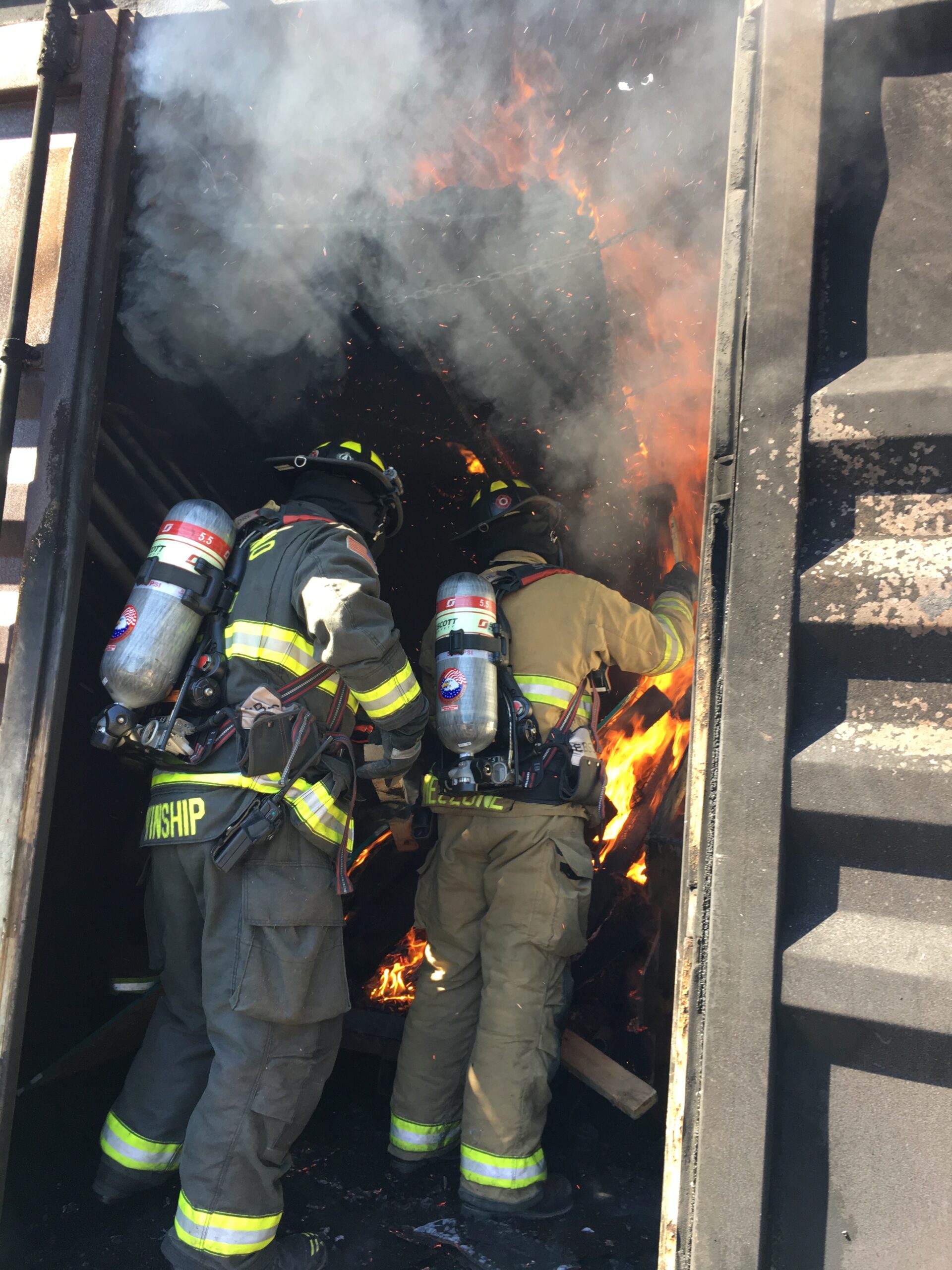 firefighters in bunker gear stand in the door of a container with flames in the background