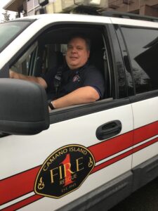 a man in blue shirt sits in the driver's seat of a white SUV with red stripe down the side and camano fire logo on the door