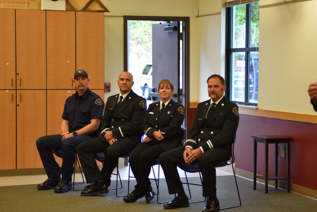 three male and one female firefighters in dress uniforms sit in chairs in a meeting room looking at a speaker
